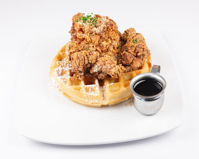 Fried chicken and waffles with powdered sugar, chives, and a side of syrup on a white plate.