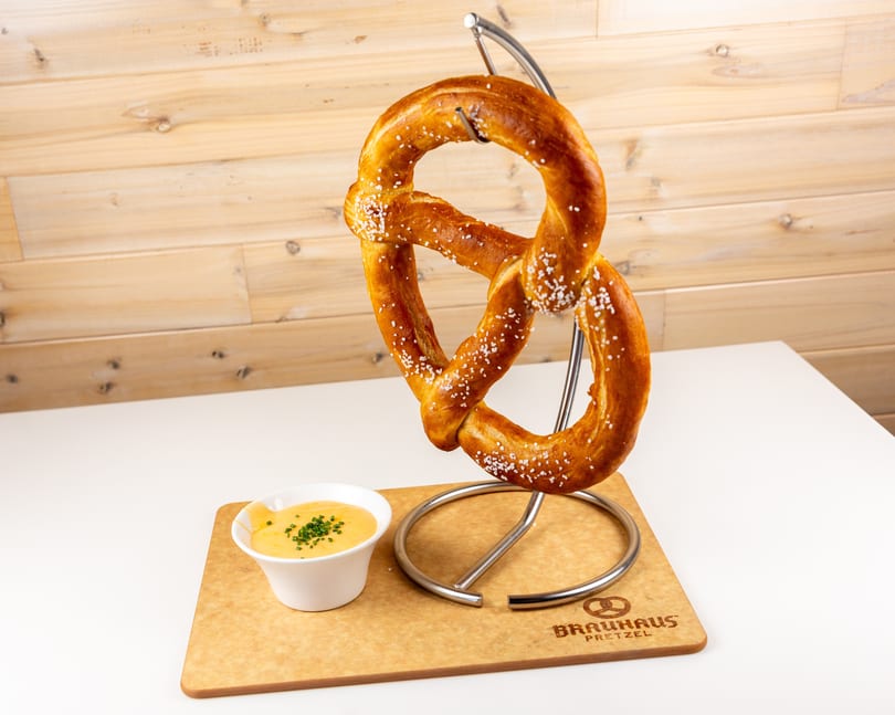 Large salted pretzel on a metal stand with a bowl of cheese dip and chives on a Brauhaus Pretzel cutting board.