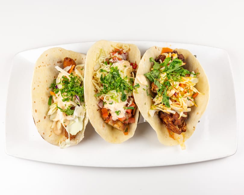 Three vibrant tacos with meat, cabbage slaw, pico de gallo, and cilantro on a white plate.
