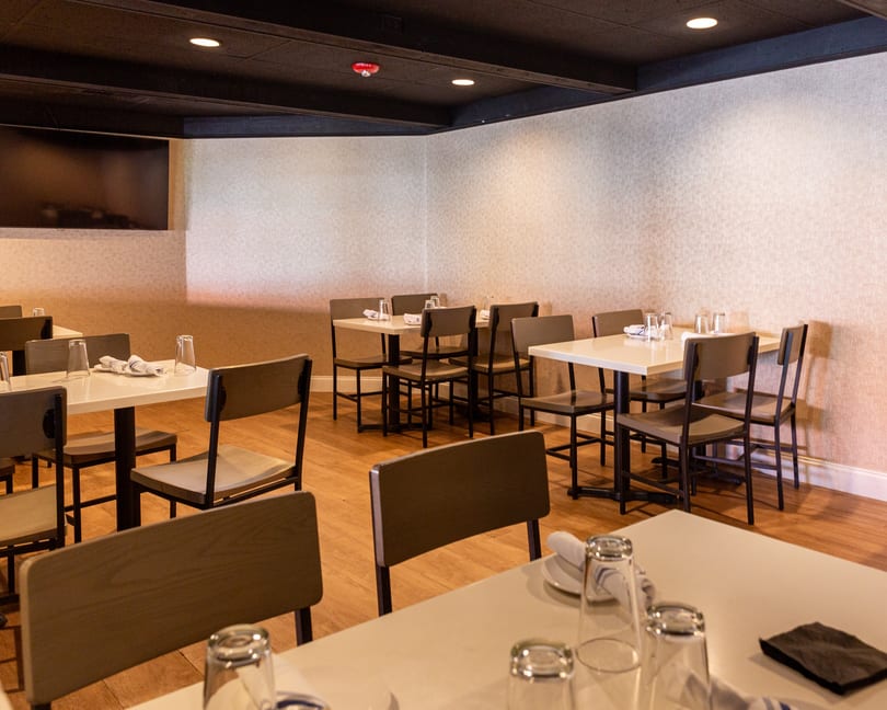 Spacious restaurant dining room with neatly set tables, chairs, and a large screen on the wall.