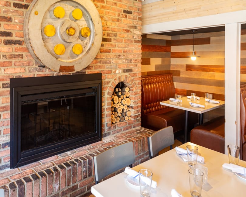 Cozy restaurant interior featuring a brick fireplace, decorative wheel, stacked wood, and set tables with brown booths.