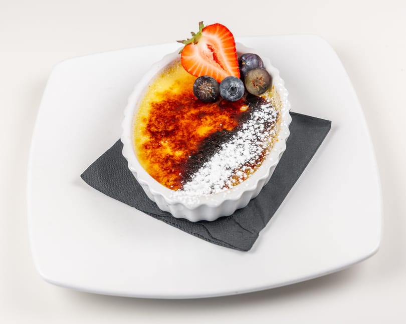 Caramelized creme brulee with powdered sugar, a strawberry half, and blueberries in a white ramekin.